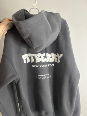 Mörkgrå hoodie från Fitberry - Mörkgrå hoodie från Fitberry med vit text på både bröstet och ryggen. Modellen har en stor huva och ribbade muddar vid ärmslut och nederkant. Materialet är mjukt. 