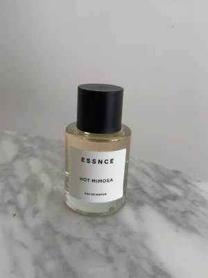 ESSNCE Hot Mimosa Eau de Parfum - ESSNCE Hot Mimosa Eau de Parfum i en stilren, rund glasflaska med svart lock. Doften har en modern och fräsch känsla, perfekt för dig som gillar unika och trendiga parfymer. Flaskan har en enkel vit etikett med svart text.