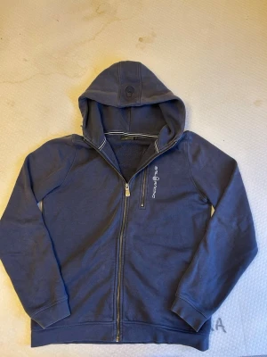 Marinblå hoodie från Sail Racing - Snygg marinblå hoodie från Sail Racing med dragkedja framtill och en extra bröstficka med zip. Broderad logga på bröstet och huva med diskret logga. Tillverkad i mjuk bomullsblandning, perfekt för chill dagar eller när du vill ha en clean look. Lite fläck på sista bilden