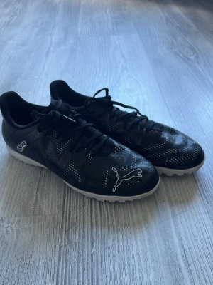 Svarta Puma fotbollsskor med vita detaljer - Snygga svarta fotbollsskor från Puma med vita detaljer och logga på tån. Skorna har snörning och en låg profil, samt små vita prickar över hela ovandelen för extra stil. Yttersulan är vit och greppvänlig, perfekt för spel på konstgräs eller grus.