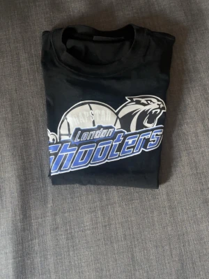 London Shooters svart t-shirt - Svart t-shirt med tryck av London Shooters och en panter samt basketboll på bröstet. T-shirten har rund hals och korta ärmar. Trycket är i vitt, blått och grått vilket ger en sportig vibe. Perfekt för dig som gillar basket eller vill ha en streetstyle-look.