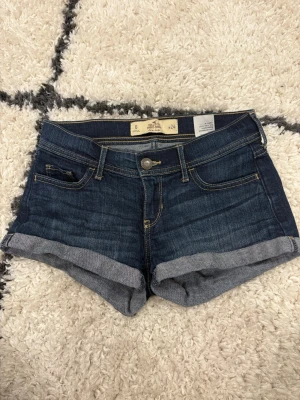 Mörkblå jeansshorts från Hollister - Snygga mörkblå denimshorts från Hollister med låg midja och uppvikta benslut. Perfekta för varma dagar och enkel att matcha med allt. Skriv om du har frågor! 