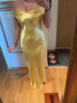 Gul axelbandslös långklänning - Elegant gul långklänning i glansigt material med offshoulder-design och figurnära passform. Klänningen har en vacker omlott-detalj över bysten och faller mjukt längs kroppen. Perfekt för sommarens speciella tillfällen.