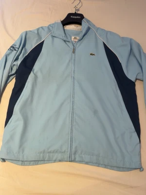 Lacoste windbreaker - En blå  lacoste  windbreaker i storlek 164 cm/14 år  med en fläck som kanske går bort i tvätten men inte jätte synlig