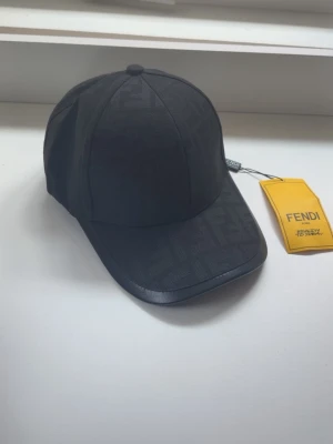 Svart keps från Fendi med logomönster - Snygg svart keps från Fendi med diskret FF-logomönster över hela kepsen. Klassisk böjd skärm och justerbar passform. Tillverkad i ett slitstarkt tyg med exklusiv känsla. Perfekt accessoar för en trendig och stilren look.