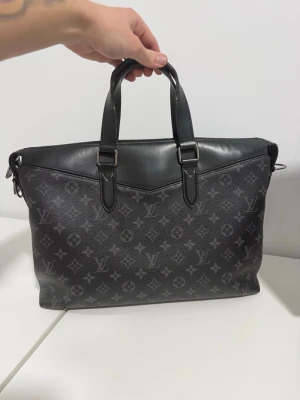 Louis Vuitton svart datorväska - Snygg svart datorväska från Louis Vuitton med klassiskt monogrammönster i grått. Väskan har två handtag, silverfärgade metalldetaljer och är tillverkad i tåligt läder och canvas. Perfekt för dig som vill ha en stilren och lyxig look till skolan eller jobbet.