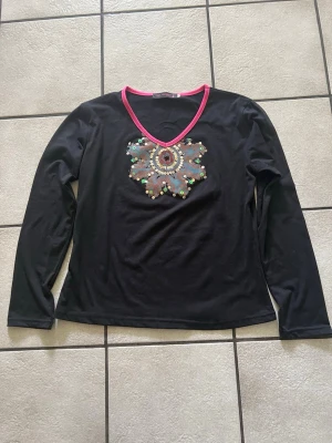 Svart topp med paljetter och blommönster storlek M/L - Svart långärmad topp från Cop. modern med v-ringning kantad i rosa. Framtill finns ett stort blommigt mönster dekorerat med paljetter i flera färger som ger extra bling. Perfekt för dig som gillar unika detaljer och färgklickar i din outfit.