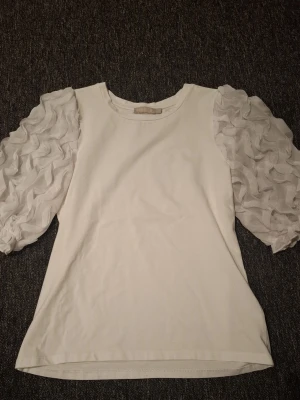 Vit topp med puffärmar från Pieces - Supersnygg vit topp från Pieces med unika puffärmar i transparent tyg och volangdetaljer. Kroppen är i mjuk bomull och ärmarna ger en extra stylish touch. Perfekt för dig som vill sticka ut lite extra med din outfit!