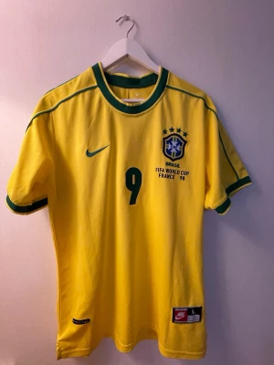 Brasilien Ronaldo 9 matchtröja Nike - Klassisk gul Brasilien fotbollströja från Nike, VM 1998, med gröna detaljer och nummer 9. Tryck med 'Ronaldo' på ryggen och officiellt landslagsmärke på bröstet. Tillverkad i lätt och ventilerande material, perfekt för fotboll eller samling.