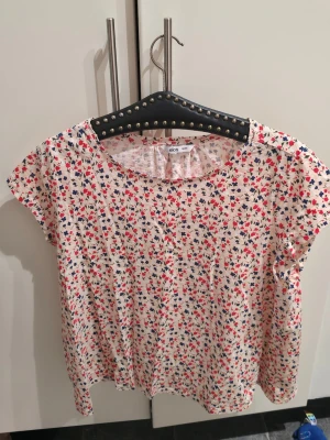 Blommig topp från Ellos - Söt blommig topp från Ellos i vitt med små röda och blå blommor. Toppen har rund halsringning och korta ärmar. Tillverkad i mjuk bomull som känns skön mot huden. Perfekt för dig som gillar färgglada och lekfulla plagg.