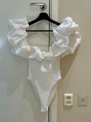  Trendig Vit body med volangkrage - Säljer en vit body med stor, dramatisk volangkrage som ger en unik och trendig look. Bodyn har hög skärning vid benen och är tillverkad i ett slätt tyg. Perfekt för dig som vill sticka ut med din outfit.