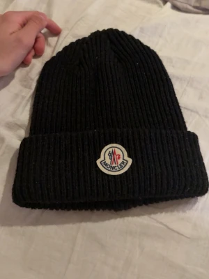 Svart Moncler ribbstickad mössa - Säljer en svart ribbstickad mössa från Moncler med uppvikt kant och klassisk Moncler-logga framtill. Mössan är i tjockt, mjukt material som håller värmen och har en stilren look som passar till det mesta.
