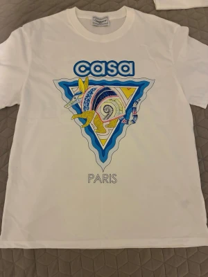 Vit Casa Paris t-shirt med tryck - Snygg vit t-shirt från Casa Paris med ett färgglatt grafiskt tryck av ett djur och texten 'casa' och 'PARIS' på framsidan. T-shirten har klassisk passform och korta ärmar. Perfekt för dig som gillar unika prints och streetstyle.