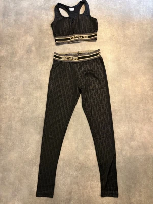 Leggings och topp - Leggings och topp med bokstäver på. Svart och guldiga. Toppen har lite ilägg som går att ta bort. Resår i midjan både på topp och byxor. Köpt som storlek S. Helt nytt!