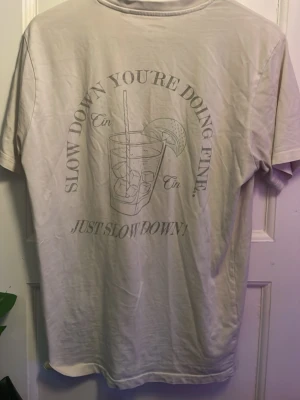 Vit t-shirt med tryck och citat - Vit t-shirt med ett stort tryck på ryggen som visar ett glas gin & tonic och texten 'Slow down you're doing fine. Just slow down!'. Klassisk passform och rund hals. Perfekt för dig som gillar chill vibes och stilrena plagg.