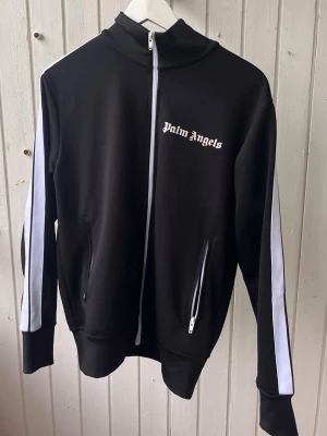 Palm Angels svart tracksuit - Säljer en svart tracksuit från  Palm Angels med vita ränder längs ärmarna och vit dragkedja. Jackan har hög krage, två fickor med dragkedja och logga på bröstet. Perfekt för en sportig och trendig streetwear-look. Helt ny