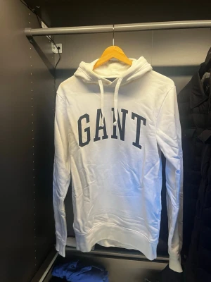 Vit hoodie från GANT med tryck - Snygg vit hoodie från GANT med stor svart logga på bröstet. Klassisk modell med huva och dragsnören, samt ribbade muddar vid ärmar och nederkant. Perfekt för en avslappnad och stilren look. Tillverkad i mjukt material för extra komfort. Finns en liten missfärgning som visas på vänster sida av luvan på bild 3, men fläcken går enkelt bort i tvätten med tvättmedel