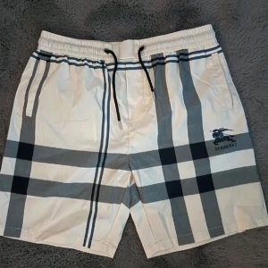 Rutig badbyxor - Snygga vita och gråa shorts från Burberry med klassiskt rutigt mönster och svart logga på benet. Shortsen har snörning i midjan och sidofickor. Perfekta för sommaren och ger en clean, trendig look.