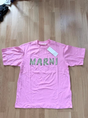 Rosa Marni t-shirt med tryck - Säljer en rosa t-shirt från Marni med grönt, handmålat tryck på bröstet. Klassisk rund hals och korta ärmar. Perfekt för dig som vill ha en statement tee från ett trendigt märke. Materialet är mjuk bomull och passformen är loose.