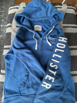 Blå zip hoodie från Hollister - Snygg blå hoodie från Hollister med dragkedja, vit text på ärmen och logga på bröstet. Hoodien har vita snören i huvan och två fickor framtill. Perfekt för en avslappnad och cool stil. Materialet är mjukt och skönt, troligen bomullsblandning.