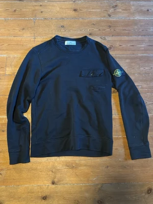 Svart sweatshirt från Stone Island - Svart sweatshirt från Stone Island med rund hals, två bröstfickor och den ikoniska logotypen på vänster ärm. Tröjan har en clean look och är tillverkad i mjukt bomullsmaterial. Perfekt för dig som gillar streetwear och vill ha något stilrent.