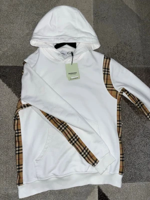 Burberry hoodie - Snygg vit hoodie från Burberry med klassiska beige rutiga detaljer längs sidorna och ärmarna. Hoodien har en stor huva, ribbade muddar och en praktisk magficka. Perfekt för dig som vill ha en stilren och trendig look med exklusiv känsla.