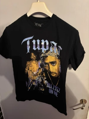 Svart Tupac t-shirt med tryck - Säljer en svart t-shirt med stort Tupac-tryck på framsidan och texten 'All Eyez On Me'. T-shirten har klassisk passform, rund hals och korta ärmar. Perfekt för dig som gillar hiphop och streetwear.