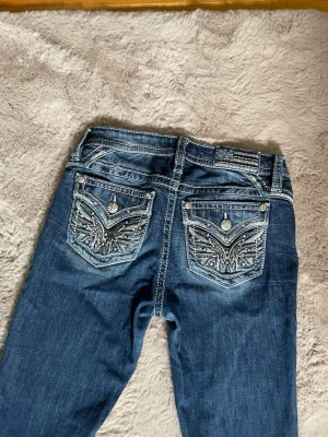 Miss Me jeans med broderade fickor - Snygga blå jeans från Miss Me med coola broderade bakfickor och dekorativa nitar. Jeansen har en klassisk femficksmodell, kontrastsömmar och en lätt tvättad look. Perfekta för dig som gillar detaljer och vill sticka ut lite extra.