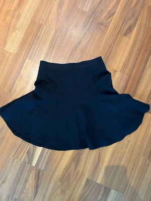 Svart kortkjol med utsvängd form - En svart kortkjol med utsvängd siluett och bred linning. Kjolen har en enkel, stilren design utan synliga detaljer eller mönster. Perfekt för att matcha med olika toppar och sneakers eller boots.