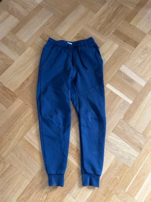 Blåa Nike tech mjukisbyxor - Säljer ett par blåa mjukisbyxor från Nike med dragsko i midjan och muddar vid bensluten. Byxorna har två sidofickor och en liten Nike-logga på vänster ben. Perfekta för chill dagar eller träning. Har även matchande hoodie på min profil:)