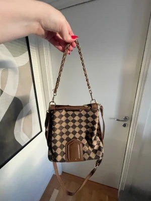 Brun axelväska med rutigt mönster - Snygg brun axelväska med klassiskt rutigt mönster i beige och mörkbrunt. Väskan har både kedjehandtag och justerbar axelrem i läderimitation. Från Shein 