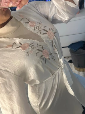 Vit sidenmorgonrock med broderi - Elegant vit morgonrock i sidenliknande material med vackra broderade blommor i ljusrosa och grönt längs kragen. Morgonrocken har knytband i midjan och vågig kant vid öppningen. Perfekt för en mysig hemmakväll.