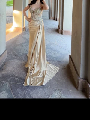 guldfärgad långklänning med paljetter - Elegant långklänning i champagnefärg med glittriga paljettdetaljer upptill och markerad midja. Klänningen har v-ringning, smala axelband och ett svepande släp. Perfekt för speciella tillfällen och bal. från oh hello clothing med alla prislapp på
