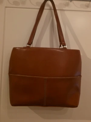 Brun läderhandväska från Kate Spade - Snygg brun handväska i äkta läder från Kate Spade New York. Väskan har två axelband, dekorativa vita sömmar och en stilren, fyrkantig form. Perfekt för dig som gillar klassisk och tidlös design med en touch av lyx.