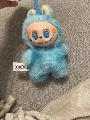 Blå fluffig BOOM nyckelring - Söt och unik blå fluffig nyckelring från BOOM med ett gulligt monsteransikte i plast och små öron. Perfekt att hänga på väskan eller nycklarna för en lekfull touch. Tillverkad i mjukt syntetmaterial och plast. Inte äkta 