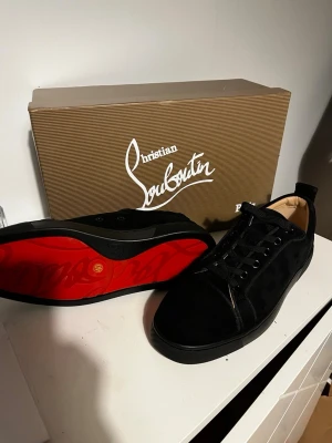 Svarta CL sneakers och plånbok - Får med grattis plånbok på köpet !!Snygga svarta sneakers från CL i mocka med klassisk röd sula och ton-i-ton broderad logga på hälen. Skorna har svarta skosnören, diskreta metalldetaljer och låg profil. Perfekta för dig som vill ha något exklusivt och stilrent.