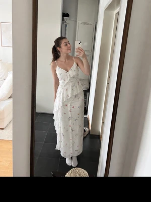 Ginatricot Vit långklänning med blommigt mönster - Romantisk vit långklänning med tunna axelband och volangdetaljer. Klänningen har ett blommigt mönster i pastellfärger och slits. Perfekt för sommarens alla tillfällen.