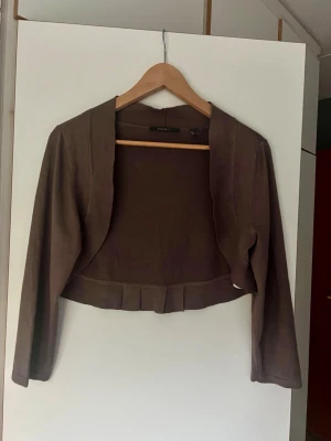 Brun bolero från Esprit  - Kort brun bolero med långa ärmar och dekorativa veck nedtill. Perfekt att slänga över en klänning eller topp för en snygg lager-på-lager-look. Enkel och stilren design som passar till mycket.