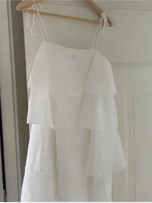Vit volangklänning från Gina Tricot - Säljer en vit klänning från Gina Tricot med flera lager av volanger och tunna axelband som knyts. Klänningen är kort och har en luftig känsla, perfekt för varma dagar. Materialet är troligtvis polyester och designen är stilren och modern.