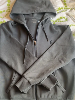 Grå hoodie från Polo Ralph Lauren - Snygg grå hoodie från Polo Ralph Lauren med dragkedja framtill och klassisk huva med dragsko. Diskret logga broderad på bröstet och två fickor framtill. Tillverkad i mjuk bomullsblandning som känns skön mot huden. Perfekt för en avslappnad och stilren look.🔥