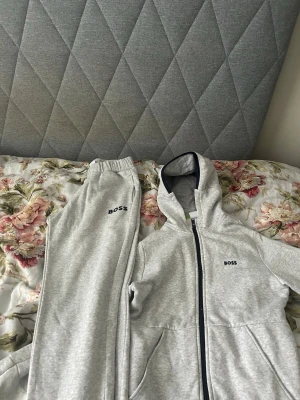Grått loungewear-set från BOSS - Säljer ett grått loungewear-set från BOSS med matchande hoodie och byxor. Hoodien har dragkedja, fickor och BOSS-logga på bröstet. Byxorna har resår i midjan och BOSS-logga på benet. Perfekt för chill dagar hemma.