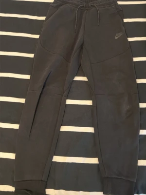 Svarta mjukisbyxor från Nike - Svarta mjukisbyxor från Nike med dragsko i midjan och ribbade muddar vid bensluten. Byxorna har fickor på sidorna och en extra ficka med dragkedja bak. Tillverkade i mjukt material för maximal komfort och avslappnad stil.