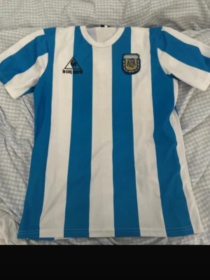 Argentina Maradona tröja Le Coq Sportif - Klassisk Argentina fotbollströja med blå och vita ränder, officiellt AFA-märke på bröstet och Le Coq Sportif-logga. På ryggen står det 'Maradona' och nummer 10 i svart. Perfekt för dig som älskar retro och fotbollshistoria.
