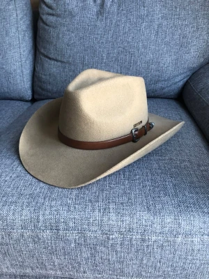 Beige cowboyhatt från Stetson - Säljer en beige cowboyhatt från Stetson i filtmaterial. Hatten har bred brätte och ett brunt läderband med snygga silverdetaljer runt kronan. Perfekt för dig som vill sticka ut med westernstil och klassisk look.