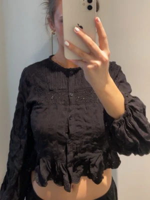 Svart blus med volangkant och brodyr - Svart blus med lång ärm, knappar framtill och volangkant nedtill. Blusen har fina broderade detaljer och små hålmönster upptill samt plissering vid bröstet. Tillverkad i ett tunt, lätt tyg som ger en luftig känsla.