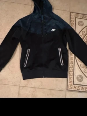Svart och mörkblå Nike hoodie - Snygg hoodie från Nike i svart och mörkblått med dragkedjeförsedda fickor och vit Nike-logga på bröstet. Hoodien har huva med dragsko och ribbade muddar vid ärmslut och nederkant. Perfekt för en sportig och avslappnad stil.
