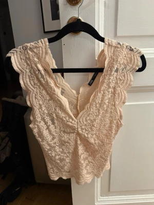 Krämrosa spets topp från Zara - Säljer en superfin krämrosa spets topp från Zara med v-ringning och scallop-kantade ärmlösa axlar. Toppen har ett romantiskt spetsmönster och är croppad, perfekt att styla med jeans eller kjol.