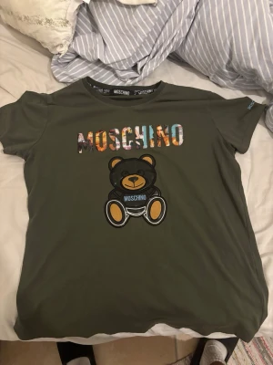 Grön Moschino t-shirt med björn - Snygg grön t-shirt från Moschino med färgglad logga och en stor björn på bröstet. Klassisk rund hals och korta ärmar. Tillverkad i mjuk bomull som känns skön mot huden. Perfekt för dig som gillar streetstyle och vill sticka ut lite extra.