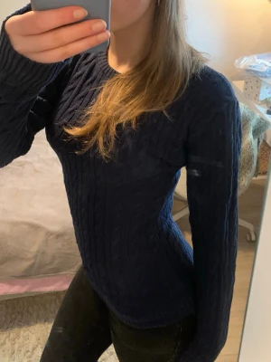 Mörkblå kabelstickad tröja - Snygg mörkblå kabelstickad tröja med rund hals och långa ärmar. Tröjan har en figurnära passform och klassiskt flätmönster. Perfekt att matcha med jeans för en stilren look.