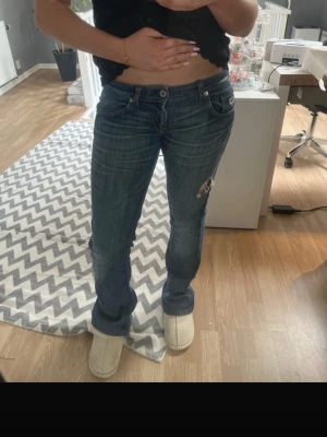 Blå bootcut jeans med slitning - Säljer ett par blå bootcut jeans med slitning och hål på ena benet. Jeansen har klassisk femficksmodell, låg midja och är tillverkade i denim. Perfekta för en avslappnad och trendig look.